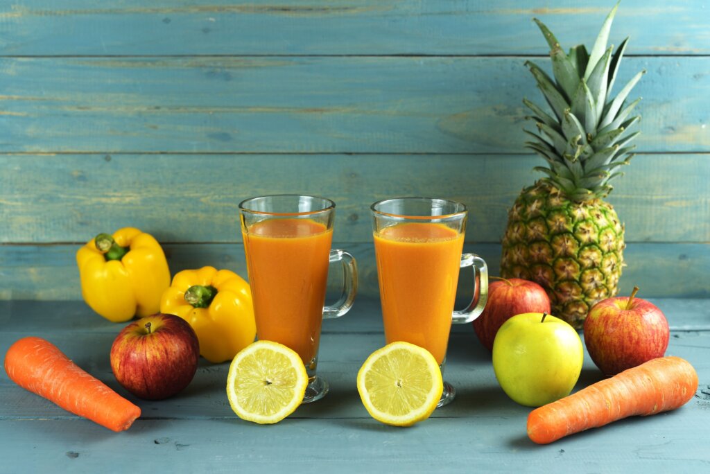 Suco detox de abacaxi, cenoura, maçã e pimentão amarelo em dois copos rodeados pelos ingredientes usados