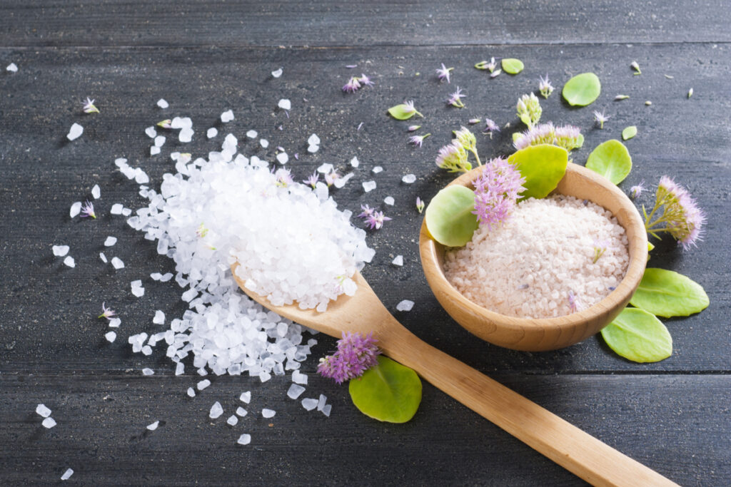 Uma colher de pau cheia de sal grosso e ao lado um pote com mais sal grosso e flores rosa