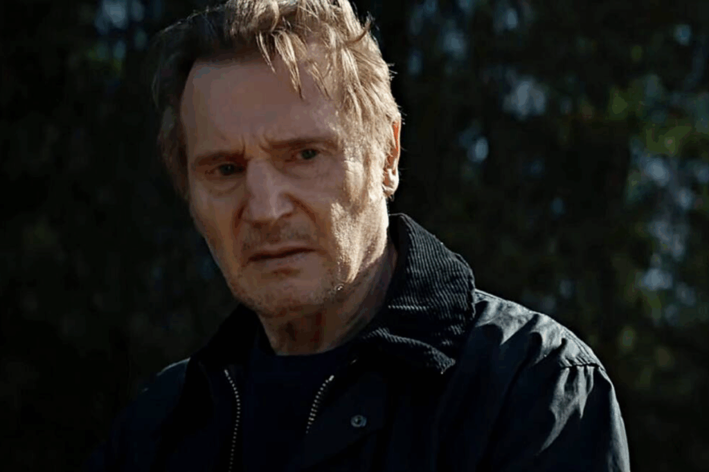 Close-up do ator Liam Neeson com uma expressão intensa e preocupada. Ele tem cabelos curtos e grisalhos, pele marcada pelo tempo e veste uma jaqueta escura com gola de veludo cotelê. O fundo está desfocado, mostrando tons escuros de verde que sugerem uma floresta ou área externa sombria.