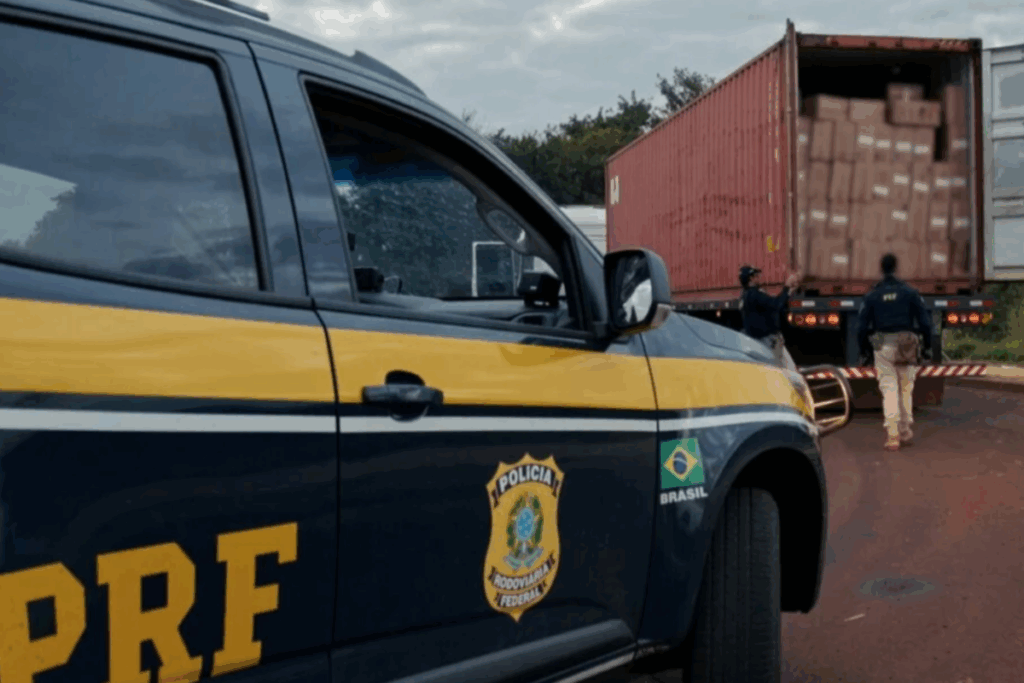 Carro preto e amarelo da PRF perto de caminhão aberto com várias caixas