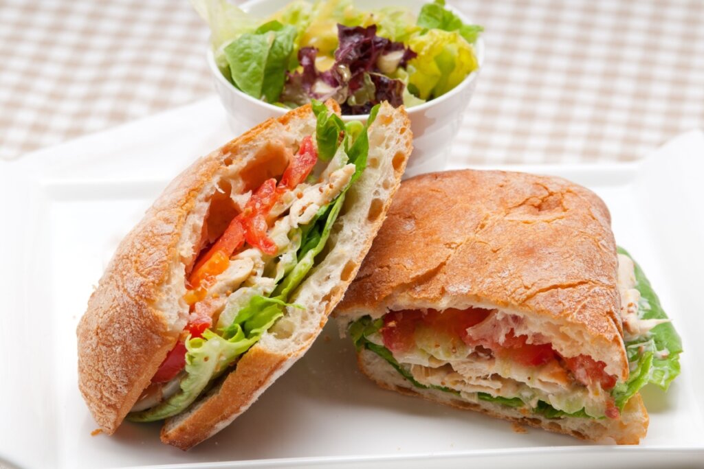 dois sanduíches de ciabatta em prato branco com pote branco de salada ao fundo
