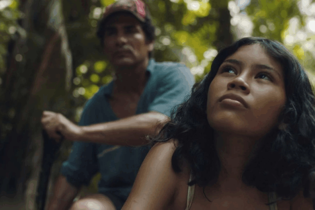 Uma jovem menina ribeirinha com cabelos escuros e ondulados, olhando para cima com uma expressão pensativa ou de expectativa. Ao fundo, ligeiramente fora de foco, um homem de boné e camisa azul segura um remo, indicando que estão em uma embarcação. O cenário é de floresta densa e iluminada pelo sol.