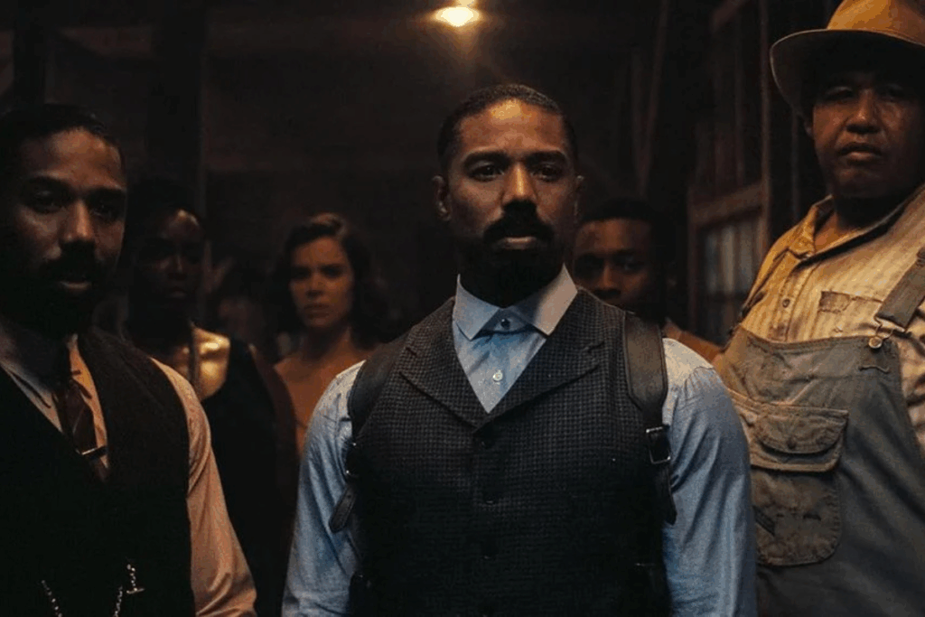 Cena do filme "Pecadores" em um ambiente rústico e sombrio. A imagem destaca dois personagens interpretados por Michael B. Jordan: um ao centro, vestindo camisa azul, colete cinza e um coldre de ombro; e outro à esquerda, com barba mais cheia, usando colete preto e gravata. Ambos mantêm expressões tensas e vigilantes. À direita, um homem robusto de chapéu e macacão completa o grupo, enquanto outras figuras aparecem ao fundo sob a luz fraca de uma lâmpada pendente.