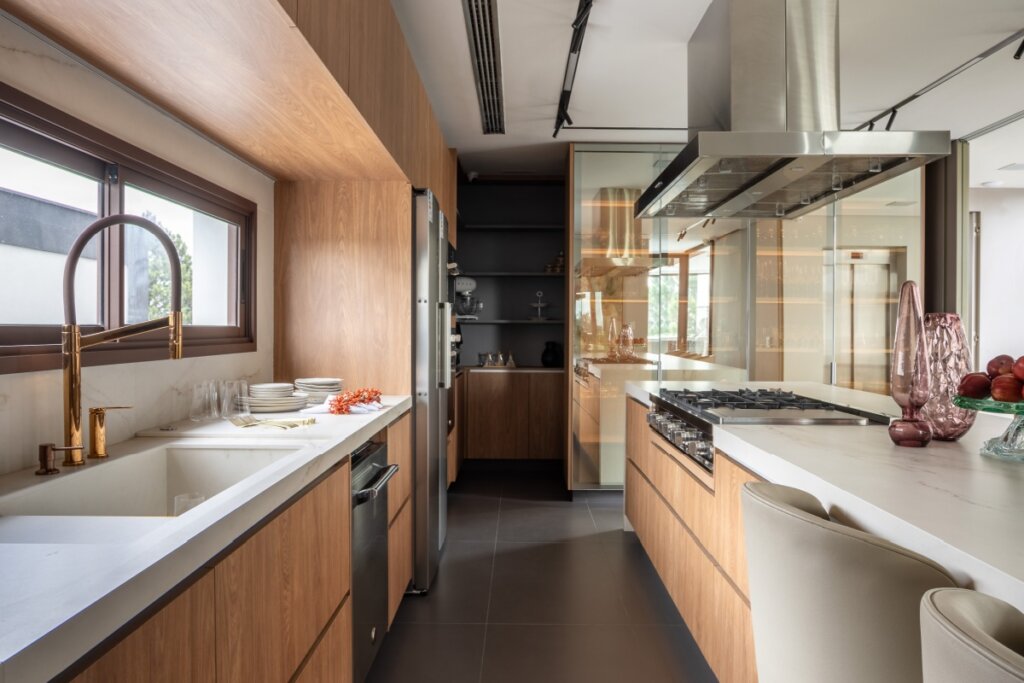Vista interna da cozinha em estilo corredor. À esquerda, bancada branca com cuba esculpida e torneira gourmet dourada. À direita, ilha com cooktop e coifa de aço inox. Ao fundo, armários com portas de vidro reflecta criam profundidade e refletem a iluminação quente do ambiente.