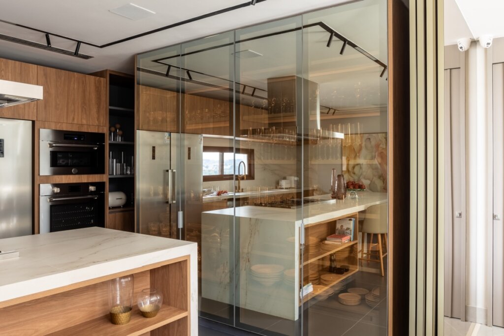 Imagem lateral da cozinha focando no fechamento de vidro reflecta que envolve a área da cristaleira e despensa. O vidro possui um tom bronzeado/champagne que permite ver levemente o interior organizado enquanto reflete a luminosidade natural da janela oposta.