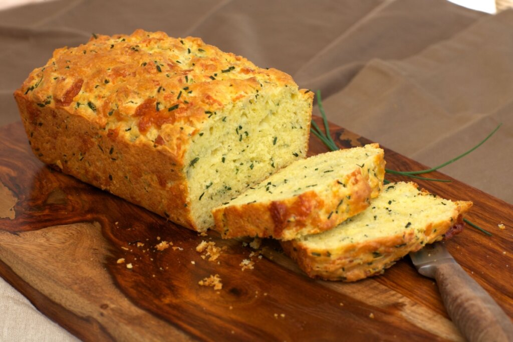 Bolo de fubá: 7 receitas práticas e deliciosas para o café da tarde 5 Bolo de fubá com queijo e cebolinha, em cima de uma tábua de madeira, com duas fatias cortadas