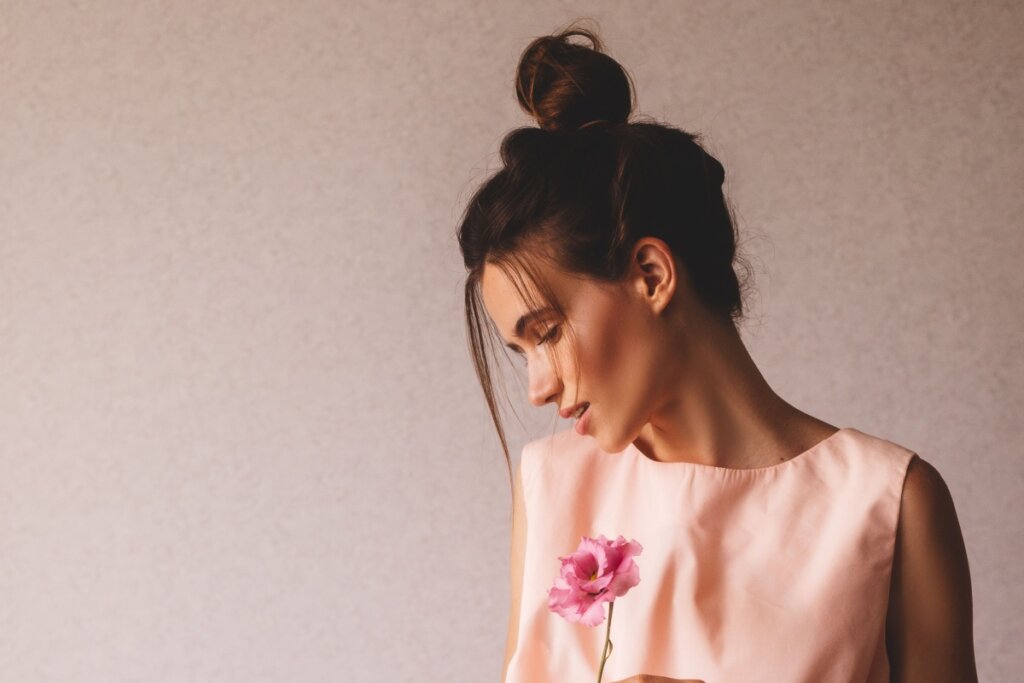 5 penteados estilosos e fáceis para curtir festivais de música 20 Fotografia de perfil de uma mulher jovem de pele clara, com o rosto levemente inclinado para baixo. Ela usa um coque alto e despojado no topo da cabeça, com duas mechas finas de cabelo soltas emoldurando o rosto. Veste uma blusa de cor rosa suave e segura uma flor rosa delicada à sua frente. O fundo é uma parede de textura simples em tom neutro, com iluminação suave vindo da lateral.