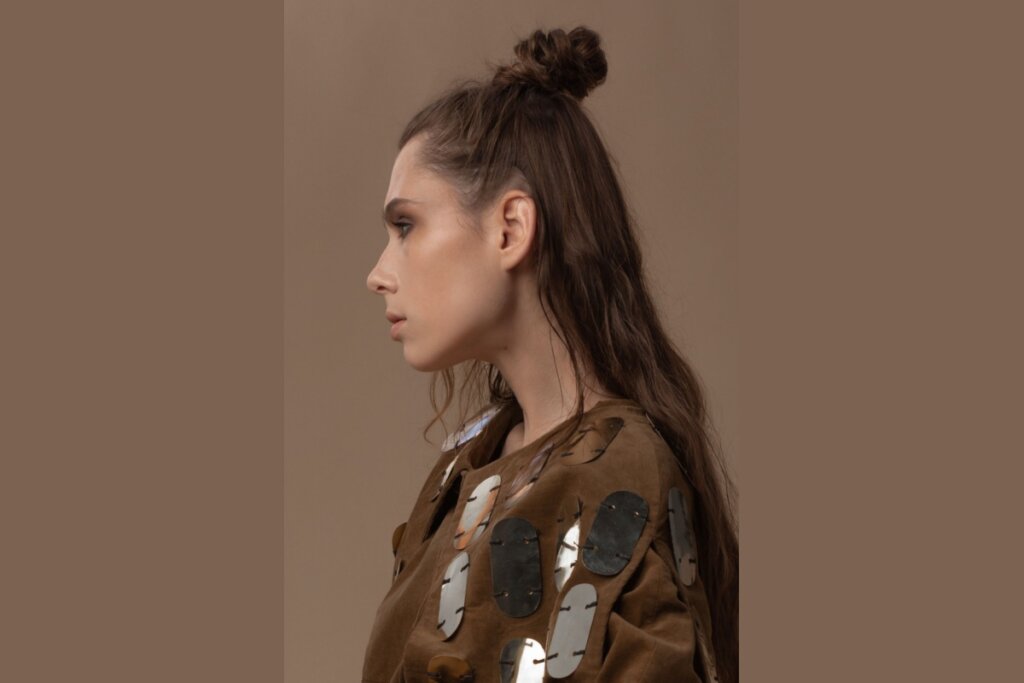 5 penteados estilosos e fáceis para curtir festivais de música 22 Fotografia de perfil de uma mulher com cabelos castanhos longos e ondulados, usando um penteado "half bun" (meio coque) alto e texturizado no topo da cabeça, com a metade inferior dos fios solta sobre as costas. Ela veste uma jaqueta marrom de camurça decorada com grandes aplicações ovais metálicas e reflexivas. A composição possui tons terrosos, com um fundo liso em bege escuro e iluminação suave que destaca o contorno do seu rosto e a textura do cabelo.