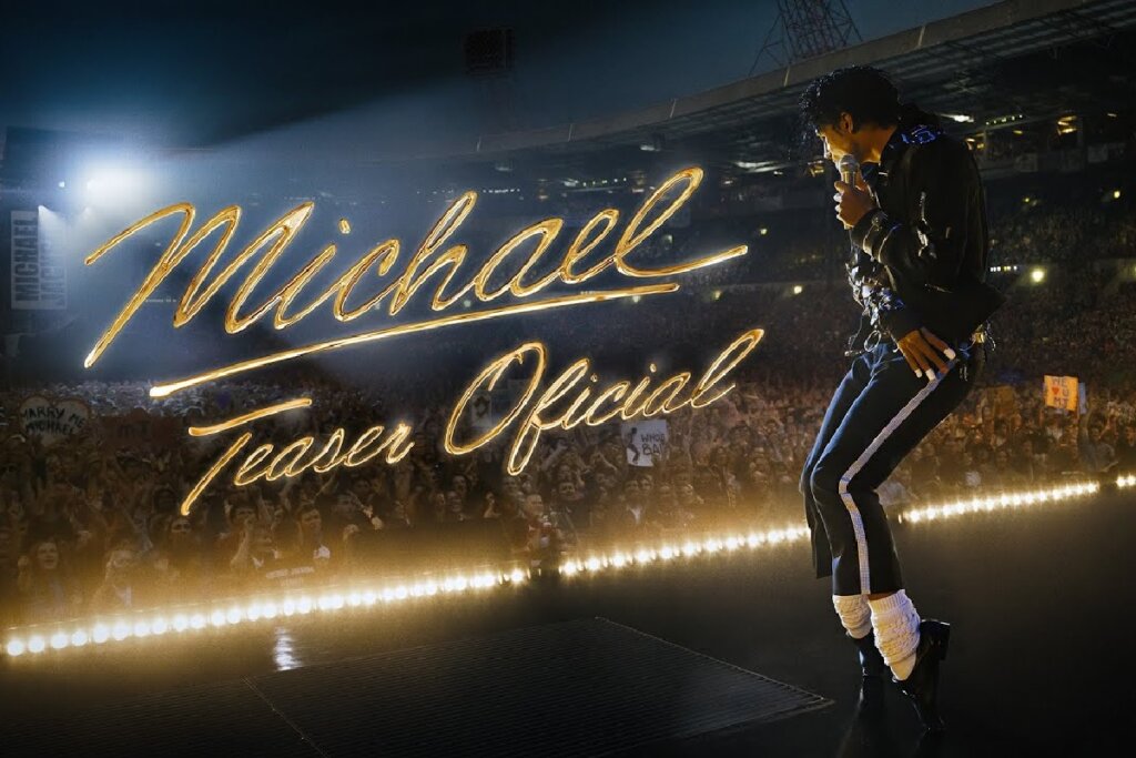 5 filmes imperdíveis que chegam ao cinema em abril 22 Imagem promocional da cinebiografia de Michael Jackson. Um ator caracterizado como o cantor está de perfil no palco de um estádio lotado, segurando um microfone. Ele usa uma jaqueta preta, calças com listras laterais brancas, meias brancas e sapatos pretos. No centro da imagem, o título "Michael Teaser Oficial" aparece escrito em letras douradas cursivas e brilhantes.