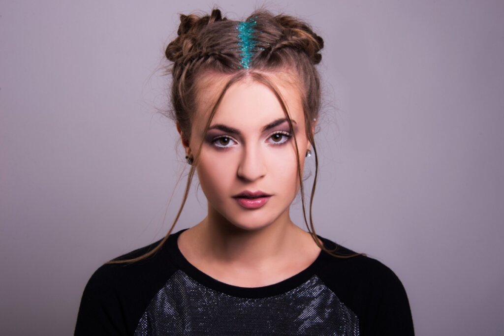 5 penteados estilosos e fáceis para curtir festivais de música 23 Retrato frontal de uma mulher jovem comO cabelo está dividido ao meio e preso em dois coques trançados nas laterais do topo da cabeça (estilo "space buns"), com a risca central da raiz preenchida por glitter azul-turquesa vibrante. Ela possui mechas finas de cabelo soltas emoldurando o rosto, usa uma maquiagem esfumada nos olhos e veste uma camiseta preta com detalhe frontal prateado. O fundo é cinza neutro e liso.
