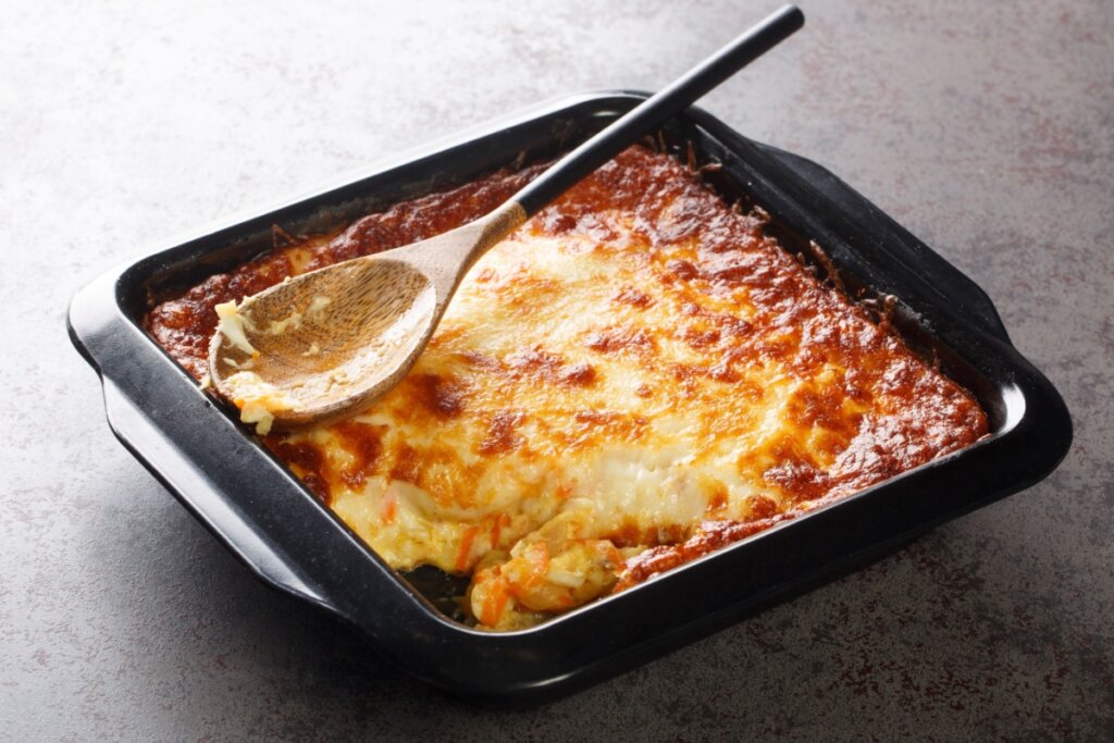 Bacalhau gratinado servido em forma preta em cima de bancada de pedra com colher de madeira em cima