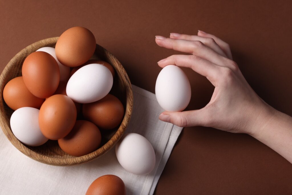 10 alimentos que não podem faltar na dieta da mulher 46 Pote de madeira com ovos brancos e vermelhos. Mão ao lado segurando ovo branco