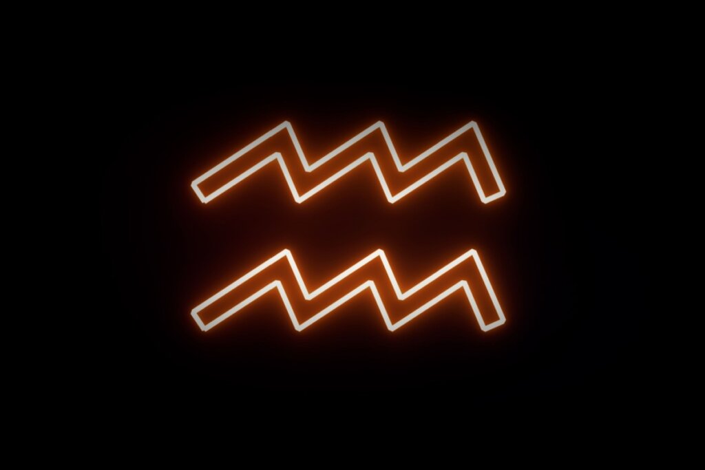 Aqui estão os textos alternativos para as duas imagens restantes, mantendo o padrão das anteriores: Aquário.jpg: Ilustração digital do símbolo de Aquário (duas linhas horizontais em zigue-zague paralelas, representando ondas de água). O símbolo possui um contorno com brilho neon em tons de laranja e branco sobre um fundo preto.