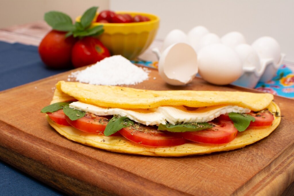 Uma crepioca dourada e dobrada ao meio está em destaque sobre uma tábua de madeira rústica. O recheio visível é composto por fatias de queijo branco, rodelas de tomate fresco, folhas de manjericão e uma pitada de ervas secas.