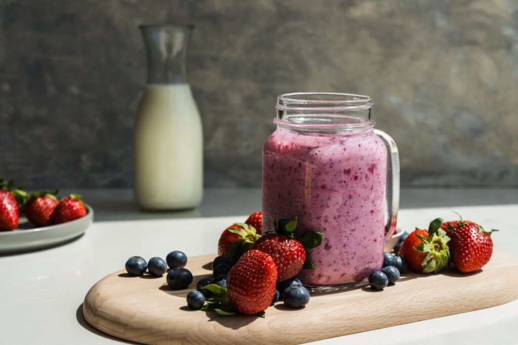 A imagem mostra uma vitamina cremosa de frutas vermelhas servida em um copo de vidro com alça. A bebida tem coloração rosada, indicando a mistura de ingredientes como morango e mirtilo.

Ao redor, aparecem frutas frescas — morangos e blueberries — além de uma jarra de leite ao fundo